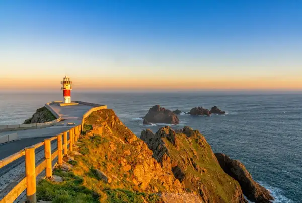Faro en el cabo Ortegal al atardecer, con el mar azul y rocas emergiendo de las olas.