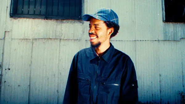 Earl Sweatshirt, smiling and wearing a blue plaid cap and dark jacket, stands against a weathered wall. He's ready to drop his next earl sweatshirt setlist.