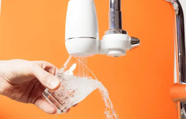 Un robinet avec un filtre blanc verse de l'eau dans un verre. C'est le moment de changer le filtre robinet pour une eau plus pure.