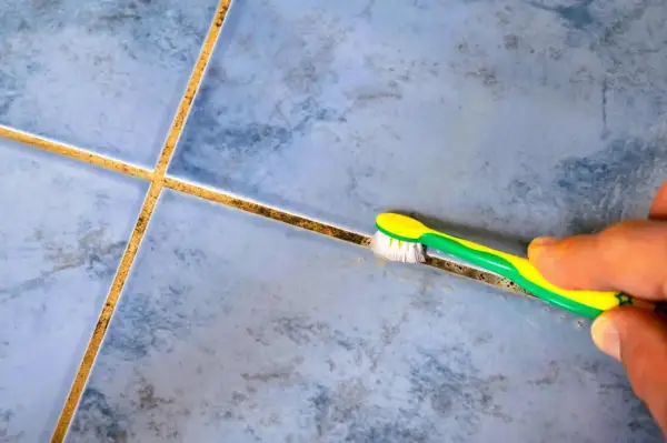 Une main utilise une brosse à dents pour blanchir le joint de carrelage sale, révélant la propreté cachée.