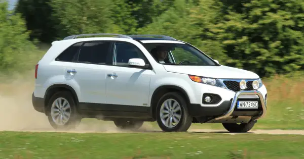 Białe Kia Sorento 1 zderza się z kurzem na drodze gruntowej, jadąc przez zielone pola.