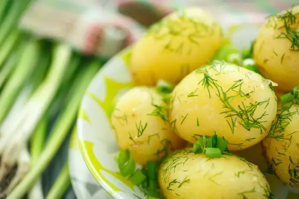 Pyszne, młode ziemniaki na parze, posypane świeżym koperkiem i szczypiorkiem, obok pęku zielonej cebulki.
