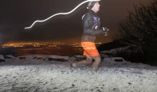 Un coureur nocturne s'entraîne pour la Saintélyon sur un sentier enneigé, la ville illuminée en arrière-plan.