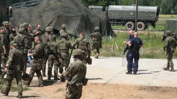Żołnierze w pełnym rynsztunku, w tym środki przymusu bezpośredniego wojsko, przygotowują się do działań. W tle wojskowe pojazdy i namioty.