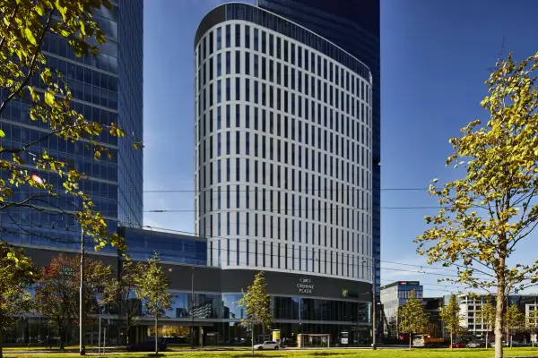 Nowoczesny budynek Crowne Plaza, część kompleksu Holiday Inn Warszawa, z charakterystyczną, falistą fasadą i zielenią wokół.