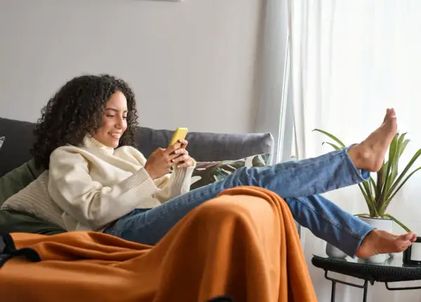 Frau mit lockigen Haaren entspannt sich richtig sitzend auf dem Sofa, schaut auf ihr Handy.