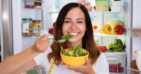 Uśmiechnięta kobieta je sałatkę z lodówki, wiedząc, że zdrowe jedzenie to klucz, jak przyspieszyć metabolizm.