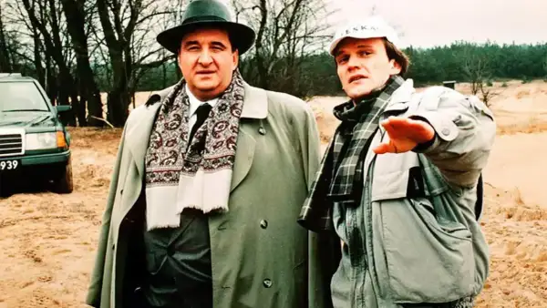 Dwóch mężczyzn w płaszczach i szalikach, jeden w kapeluszu, drugi w czapce. W tle drzewa i piasek. Nic śmiesznego.