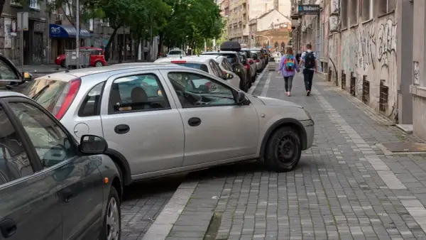 Samochody na chodniku utrudniają przejście. Dwie osoby idą wąskim chodnikiem, omijając przeszkody.