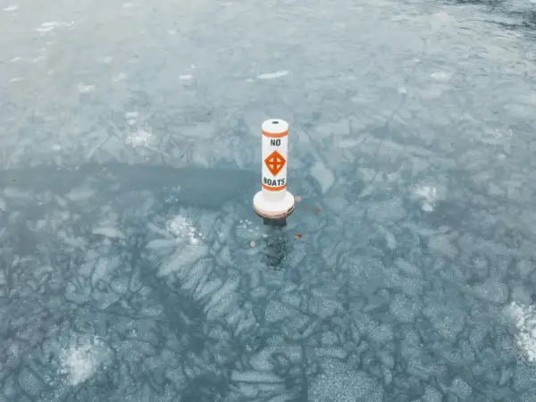 Bojka z napisem "NO BOATS" stoi na zamarzniętej tafli wody, ostrzegając przed ruchem łodzi.