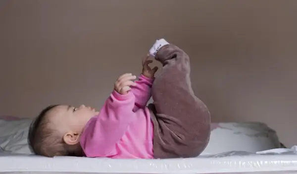 Niemowlę w różowym body i brązowych spodenkach leży na plecach, podnosząc nóżki. Pozycja ta może być pomocna w diagnostyce dysplazji stawu biodrowego.