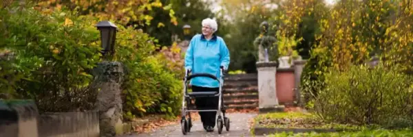 Elderly woman in blue jacket walks with a walker in a park with autumn foliage.