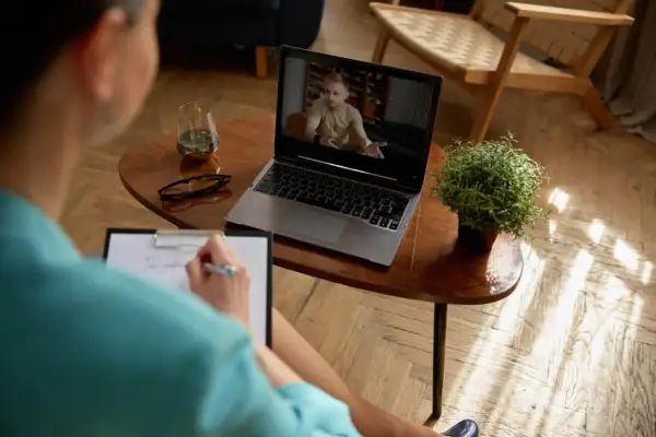 Kobieta prowadzi psychoterapię przez Skype, notując na bieżąco. Na ekranie laptopa widać mężczyznę w gabinecie.