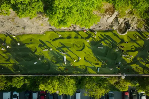 Folk spelar minigolf i Nacka Strand. En grön bana med hinder och skuggor från träden.