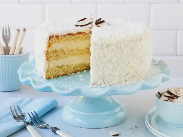 Eine köstliche Kokos Torte, bedeckt mit Kokosraspeln und dekoriert mit Kokoschips, wartet auf dem hellblauen Tortenständer.