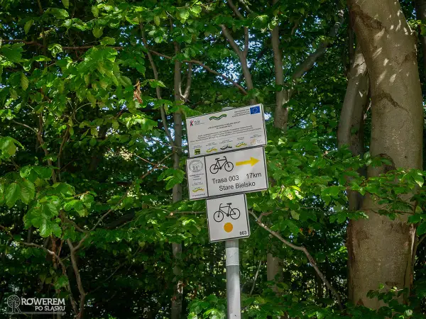 Znaki szlaku rowerowego wskazują trasę 003 do Starego Bielska, 17 km. W tle zielone drzewa.