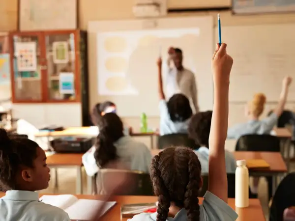Estudiantes levantan la mano en clase. El profesor proyecta información, quizás sobre el calendario escolar 2026-27.