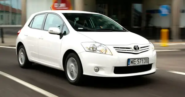 Biała Toyota Auris w ruchu miejskim, z widocznym kierowcą za kierownicą.