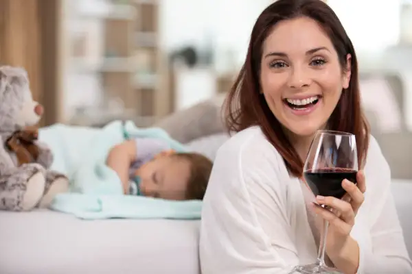 Uśmiechnięta kobieta z kieliszkiem czerwonego wina, podczas gdy dziecko śpi obok. Temat: karmienie piersią a alkohol.