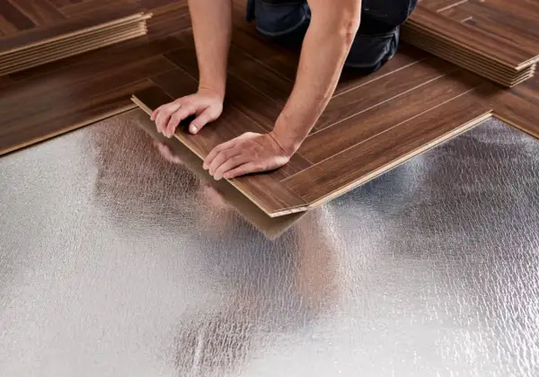 Pose bâton rompu : des mains ajustent une lame de parquet sur une sous-couche isolante argentée.
