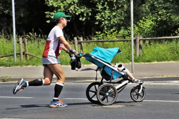 Mężczyzna w zielonej czapce i sportowym stroju bierze udział w biegu z wózkiem. Dziecko w wózku ma na sobie białe skarpetki i sandałki.