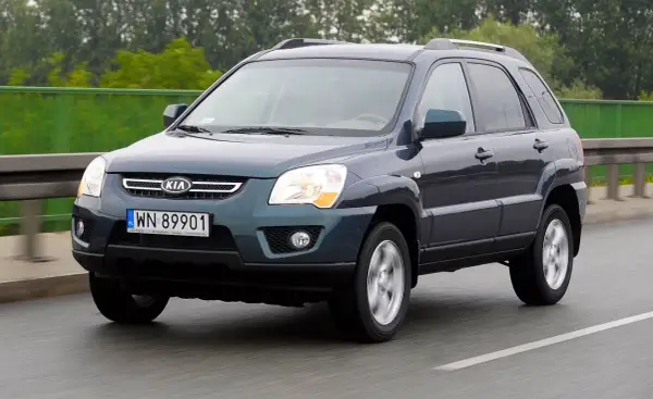 Ciemnoszary Kia Sportage 2008 na drodze, z zielonym ogrodzeniem w tle.