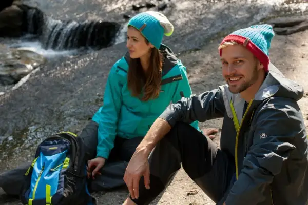 Para, co to jest trekking? Para w czapkach i kurtkach siedzi nad strumieniem, obok plecak.