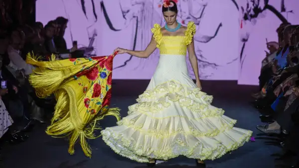 Modelo desfila en la pasarela We Love Flamenco con un vestido blanco de lunares amarillos y volantes, portando un mantón amarillo con flores rojas.
