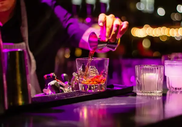 Barman nalewa bursztynowy cordial drink do szklanki z lodem. W tle kolorowe światła.