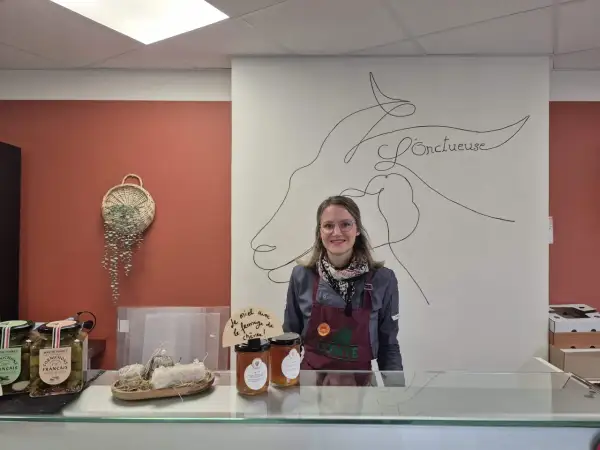 Une vendeuse souriante derrière un comptoir, avec des cornichons, du miel et du fromage de chèvre, peut-être du fromage Saint-Aubin, en vente.