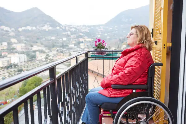 Donna in carrozzina su un balcone, con vista su un paesaggio collinare. Un'immagine che evoca la necessità di un piano nazionale non autosufficienza.
