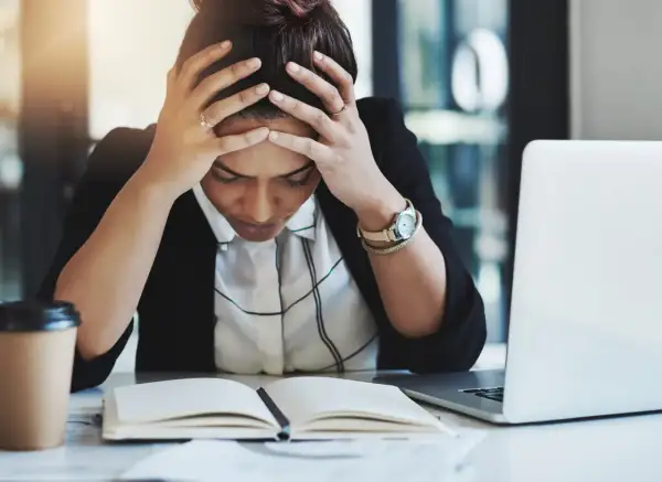 Kvinna med händerna i håret, blicken sänkt över en öppen bok. Känslan av stress på jobbet är påtaglig. En kaffekopp och laptop finns i närheten.