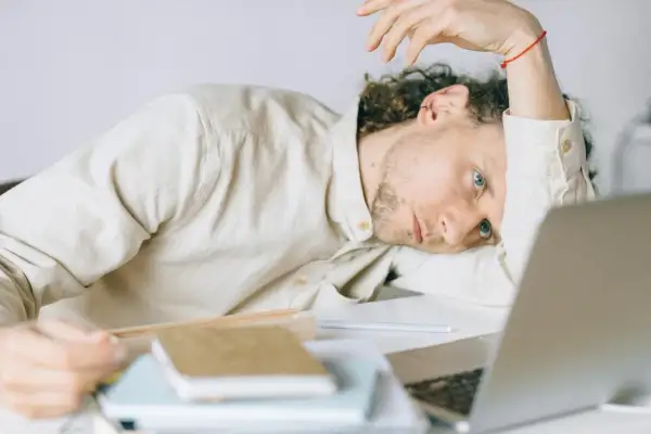 Giovane uomo con astenia neurologica, appoggiato alla scrivania, sguardo perso nel vuoto, laptop aperto.