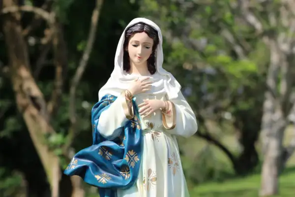 Figurka Matki Boskiej w błękitnym płaszczu ze złotymi zdobieniami. W tle drzewa. Idealna na 3 maja święto kościelne.