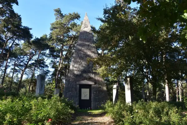 Kamienna piramida Łakińskiego w lesie, otoczona drzewami i zielenią. Obok stoją kamienne pomniki.