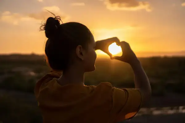 Silueta de mujer haciendo un corazón con las manos al atardecer, reflejando su actitud positiva ante la vida.