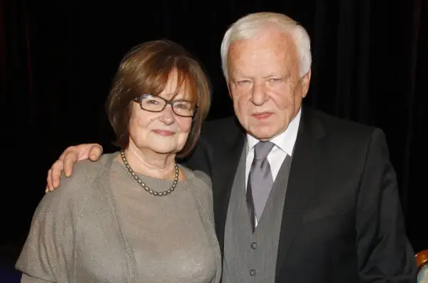 Anna Opani w okularach i z perłami na szyi, obok elegancko ubranego mężczyzny w garniturze.
