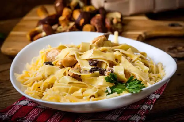 En skål med krämig pasta med kyckling och svamp, toppad med riven ost och en persiljekvist. I bakgrunden syns färska karljohanssvampar. En härlig pasta porcini-rätt.
