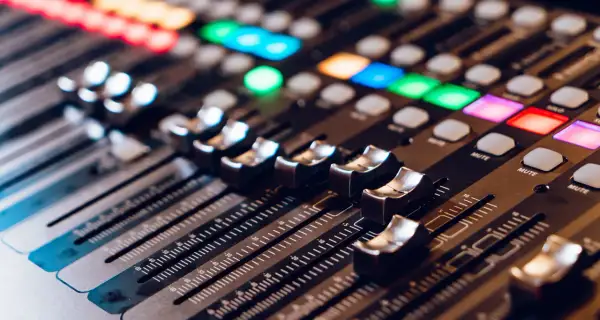 Primer plano de una mesa de mezclas con faders y botones iluminados. La **técnica de sonido** se manifiesta en este panel de control para crear la atmósfera perfecta.