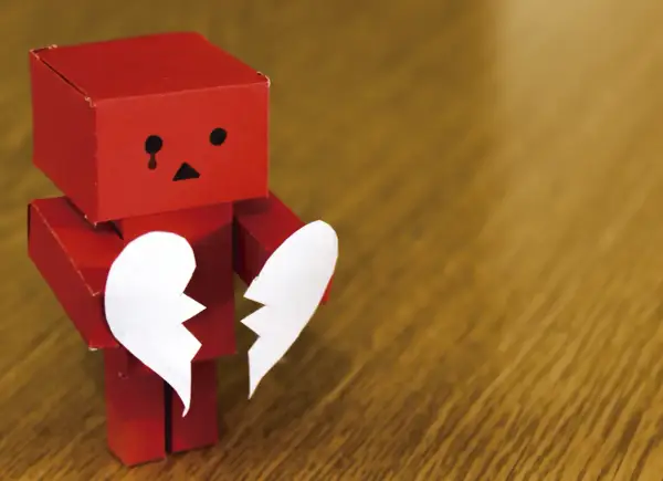 Un robot rojo de origami, con cara triste y una lágrima, sostiene las dos mitades de un corazón blanco. Parece estar a punto de morir de amor.