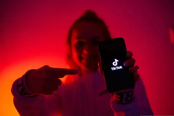 Dłoń wskazuje na ekran telefonu z logo TikToka. W tle czerwone, neonowe światła, tworzące atmosferę jak z klubu z tantiemami.