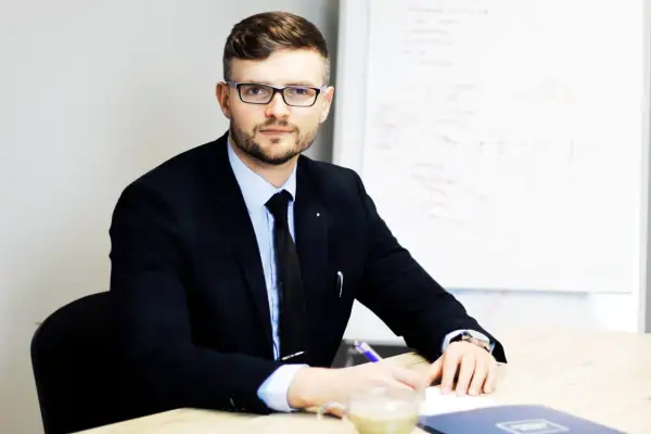 Mężczyzna w garniturze i okularach siedzi przy biurku, pisząc coś na kartce. Obok filiżanka kawy.