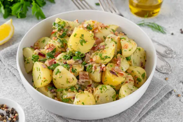 Pyszny kartoffelsalat przepis: ziemniaki z boczkiem, cebulką i ziołami w białej misce, obok cytryna i oliwa.
