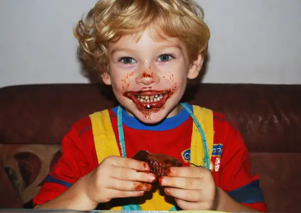 Bambino con cioccolato in bocca e sulle mani, che tiene un pezzo di torta.
