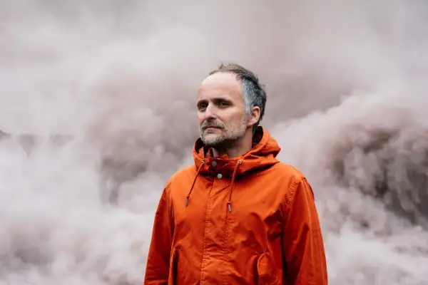 Artur Rojek w pomarańczowej kurtce, na tle dymu. Jego twórczość często porusza tematykę przemijania i refleksji.