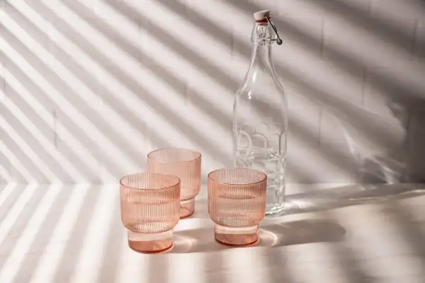 Natural product photography: three pink ribbed glasses filled with water and a clear glass bottle with a swing top, bathed in dappled light.