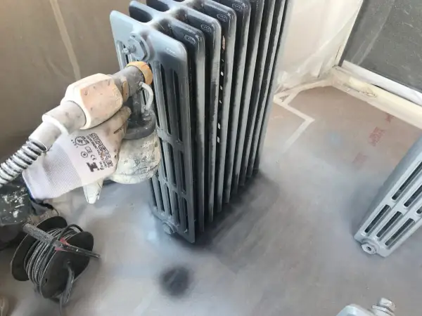 Proceso de pintar radiadores de hierro con pistola. Guante protege mano mientras se aplica pintura gris.