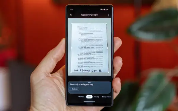 Ręka trzyma smartfon, na ekranie którego działa skaner dokumentów. Aplikacja Obiektyw Google skanuje tekst z papierowego dokumentu.