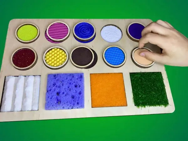 Tabla sensorial con diversos tipos de texturas para niños: círculos con materiales variados y cuadrados con esponja, fieltro y césped artificial.