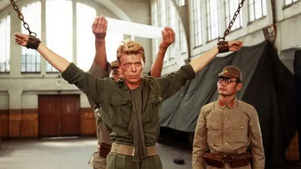 David Bowie jako major Celliers, pojmany w "Merry Christmas Mr. Lawrence", z rękami przykutymi do łańcuchów. Obok stoi japoński oficer.